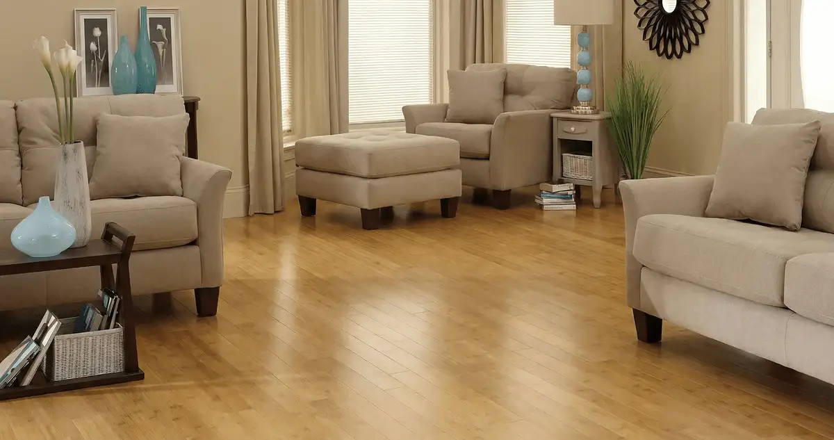 A living room with professionally installed bamboo flooring, beige sofas, a matching ottoman, a side table with a blue lamp, vases, framed pictures, and large windows with blinds and curtains in Charleston, SC
